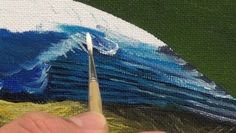 a hand holding a paintbrush in front of a painting with blue and white waves on it