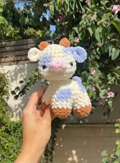 a hand holding a small crocheted cow in front of some bushes and flowers