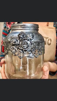 a person holding a glass jar with silver flowers on it and a tote bag in the background