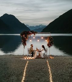 two people sitting at a table with candles in front of them on the shore of a lake