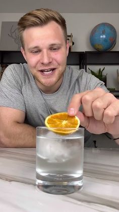 a man holding an orange slice in front of a glass filled with water on top of a table