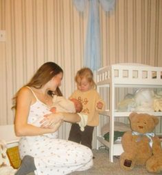 a woman holding a baby next to a teddy bear in a room with striped walls