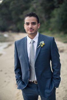 a man in a suit and tie standing on the beach with his hands in his pockets