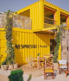 a yellow shipping container with tables and chairs on the beach next to it that reads, necpresso place