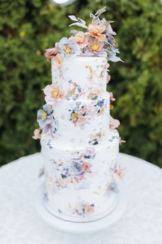 a white wedding cake with flowers on the top and bottom tier is sitting on a table in front of some bushes