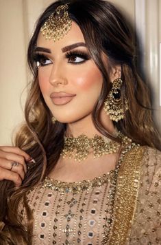 a woman with long brown hair wearing a gold dress and jewelry on her head, standing in front of a white wall