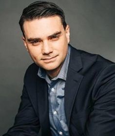 a man in a black jacket and blue shirt posing for the camera with his hands on his knees