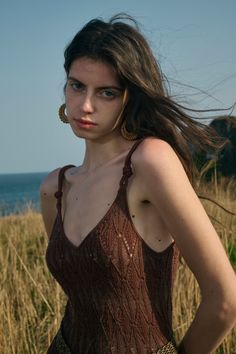 a woman with long hair standing in front of the ocean wearing a brown dress and gold hoop earrings