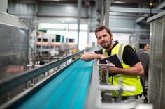 a man in a yellow vest is standing near a conveyor belt and looking at the camera