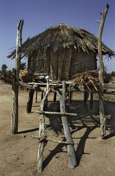 a hut made out of sticks and wood in the middle of dirt field with blue sky