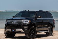 a black ford expedition vehicle parked on the beach next to water and bridge in the background