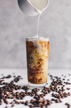 iced coffee being poured into a glass filled with ice and coffee beans on a white surface