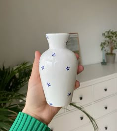 a hand holding a white vase with blue flowers on the outside and inside, in front of a dresser