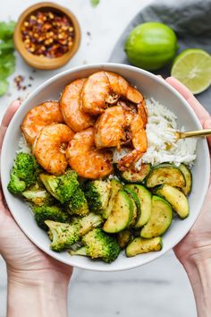 two hands holding a white bowl filled with shrimp, broccoli and cucumbers