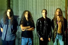 four men standing next to each other in front of a metal wall with their arms crossed