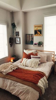 a bed with white sheets and orange blankets in a small room next to two windows