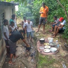 a group of people standing around cooking food on top of a wooden table in the woods