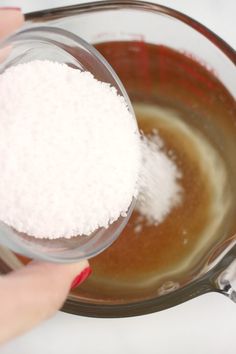 a person holding a glass bowl filled with white sugar and brown liquid next to a spoon