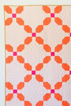 an orange and white quilt with pink squares on the top is hanging from a wall