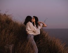 two women standing next to each other on top of a grass covered hillside near the ocean