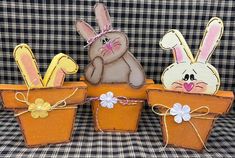 three wooden rabbits sitting in flower pots on a plaid tableclothed surface, one is wearing a pink bow and the other has an orange basket with two bunnies