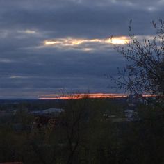 the sun is setting over some trees and buildings in the distance, with dark clouds overhead