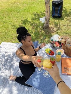 two people sitting on a blanket at a picnic table with drinks and fruit in bowls