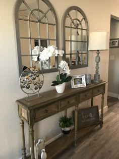 two mirrors on the wall above a wooden table with flowers and potted plants in it