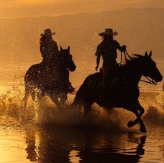 two people riding horses through the water at sunset