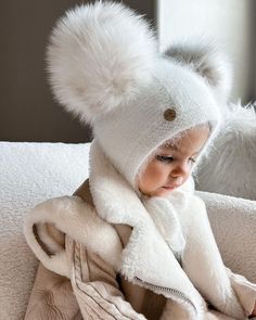 a small child wearing a white hat and scarf on top of a couch next to a teddy bear