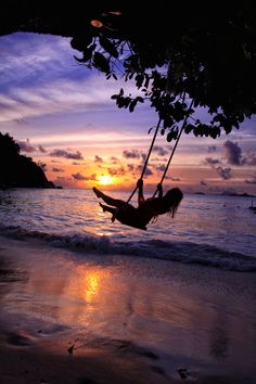 a woman is swinging on a swing at the beach as the sun sets in the background