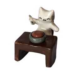 a cat figurine sitting on top of a small table with a bowl and spoon