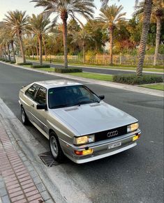 an old silver car parked on the side of the road next to some palm trees