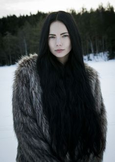 a woman with long black hair wearing a fur coat and standing in the snow near trees