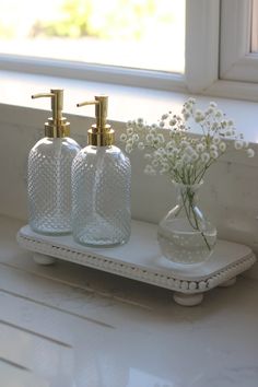 two soap dispensers on a white tray with flowers in front of a window