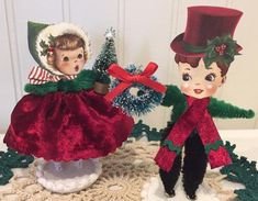 two vintage christmas dolls sitting next to each other on top of a doily covered table