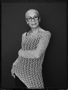 an older woman wearing glasses leaning against a wall with her arms crossed and looking at the camera