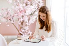 a woman sitting at a white table writing on a tablet with a cup of coffee in front of her