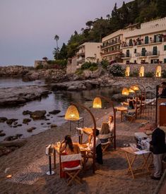 people sitting at tables on the beach next to an ocean side restaurant with lights on