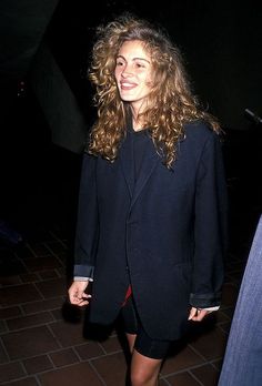 a woman with long curly hair wearing a black blazer and red skirt is smiling at the camera