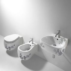 two toilets and a sink in a white bathroom with floral designs on the side wall