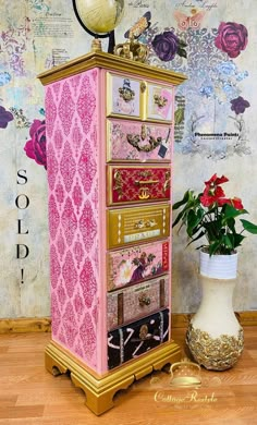 a pink and gold jewelry cabinet next to a vase with flowers in it on a wooden floor