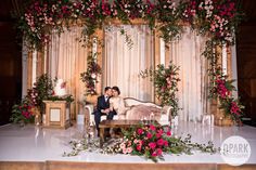 a bride and groom standing in front of a stage with flowers on the wall behind them