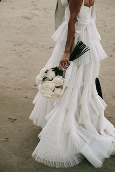 a woman is walking on the beach in a white dress and holding a bouquet of flowers