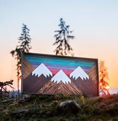 a wooden sign with mountains painted on it in front of some trees and grass at sunset