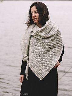 a woman standing next to the water wearing a shawl and holding her hands in her pockets