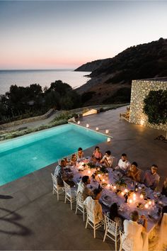 a group of people sitting around a table next to a swimming pool at night with candles on it