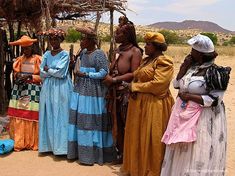 several women dressed in traditional african clothing stand together under a shelter on the desert floor