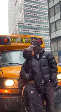 a man and woman standing in front of a school bus on a city street with snow falling all around them