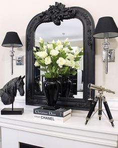 a black vase with white flowers in it sitting on a mantle next to a mirror